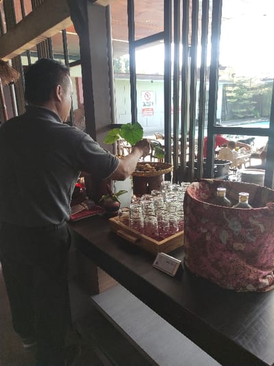 A man, wearing a black t-shirt and black pants, stands facing a table, his back to the camera. He is holding a branch of green leaves with his right hand. He is in the process of arranging glass cups in a wooden tray. The tray is filled with empty glass cups, and a large decorative container covered with a patterned fabric is in the upper right corner. The table and the floor are covered in brown wood laminate. Behind the man, a window with a white wall is partially visible. There is a "No Smoking" sign on the wall. This scene is likely a cafe or restaurant. The coordinates provided, -8.434 latitude, 114.18 longitude, are for the city of Gambiran, Indonesia.