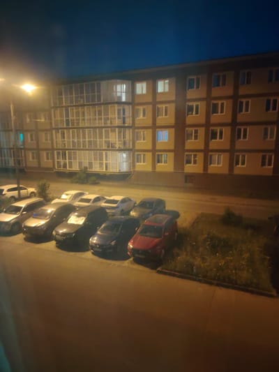 The image captures a nighttime view of a residential building and a row of parked cars in Сухарево, Russia. The building, with multiple floors and balconies, is illuminated by artificial lights, and its windows show varying degrees of light within. A streetlamp casts a bright, warm glow on the left side of the scene. In front of the building, a line of cars is parked along a street or parking area. The vehicles appear to be of various makes and models, and some are partially obscured by shadows. The ground is paved, and there is a patch of grass and vegetation to the right of the parked cars. The sky is dark, indicating it is nighttime.