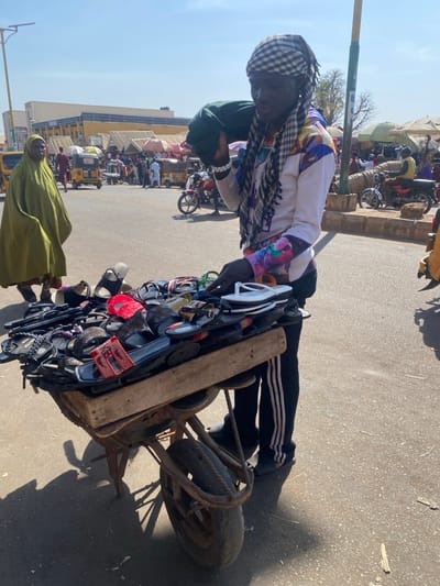 An outdoor scene in Sabon Gari South, Nigeria, depicts a male street vendor standing beside a single-wheeled wheelbarrow modified with a wooden top to display numerous pairs of sandals and slippers in various colors, including red, black, and white. The vendor, of dark complexion, wears a white long-sleeve shirt with colorful patterned lower sleeves, black track pants with three white stripes, and dark sandals. A checkered white and black scarf is wrapped around his head, and a folded green garment rests on his right shoulder. His left hand is positioned on the merchandise. In the mid-ground to the left, an adult female in an olive-green robe walks away from the viewer. The background reveals a bustling street environment with multiple individuals, yellow auto-rickshaws, motorcycles, and market stalls with awnings, all under a clear, bright sky.