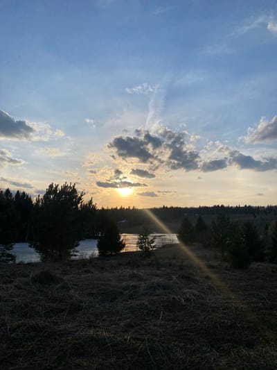 The image presents a landscape at what appears to be dusk. The sky is predominantly blue, interspersed with light and dark clouds, some obscuring a setting sun. A forest line bisects the image horizontally. Below, there is what could be an iced-over body of water and some vegetation. A ray of sunlight cuts across the foliage. Based on the location provided, this scene is likely in Ореховая Гора, Russia. No people, specific subjects, objects, actions, or interactions are clearly discernible. The primary focus is on the natural environment.