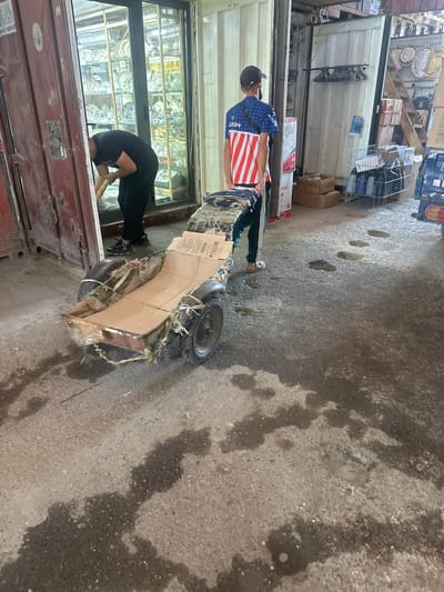 In the city of Kara-Suu, Kyrgyzstan, a scene unfolds in an open-air market or storage area. Two men are present. One man, wearing a black t-shirt and dark pants, is bent over, appearing to work near a red shipping container. The other man, dressed in a blue t-shirt with a red and white striped pattern and dark pants, is standing and pushing a makeshift cart. This cart, constructed from what appears to be discarded materials like cardboard and old tires, is loaded with fabric or textiles. The ground is a rough asphalt surface, showing significant wet patches and puddles, suggesting recent rain or a spill. In the background, more shipping containers and open market stalls filled with goods, possibly ceramics and home decor, are visible. The environment is industrial and commercial, with a mix of raw materials and finished products.