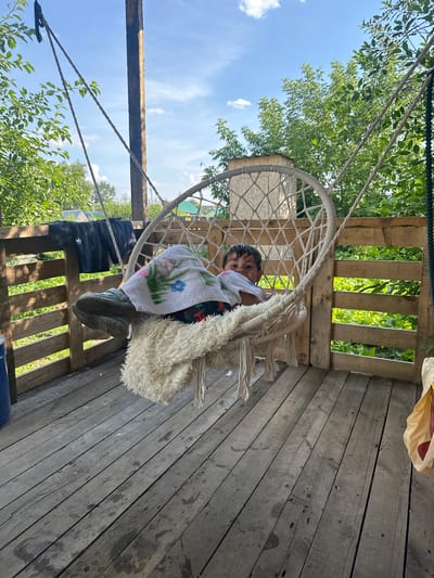 A child is lounging in a white macrame swing chair on a wooden porch. The child is covered with a towel and a fluffy white blanket. The porch overlooks a rural area with lush green trees and vegetation. A wooden fence runs along the back of the porch. The scene appears to be in a residential area of Shchuchinsk, Kazakhstan, given the style of the houses and the surrounding vegetation. The weather is sunny with a few clouds in the sky.