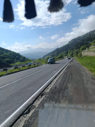 This media file captures a scenic view of a mountain road in Semyonovka, Armenia. The image is taken from a moving vehicle, looking forward and slightly to the right. The foreground shows a section of the asphalt road with a white line marking its edge. The road itself appears somewhat worn, with visible cracks and a damaged shoulder on the right side.

In the mid-ground, several vehicles are traveling on the road. A dark blue van is prominently positioned in the left lane, while a black car is further ahead in the right lane. Beyond these vehicles, the road curves into the distance, bordered by lush green hills and dense forests that climb the steep mountain slopes. A black metal guardrail with alternating white and black reflective stripes runs along the left side of the road.

The background is dominated by majestic mountain ranges that fade into a hazy blue, suggesting a considerable distance. The sky is a vibrant blue, dotted with fluffy white clouds, indicative of pleasant weather, likely midday given the bright sunlight casting shadows. A few utility poles with power lines are visible against the mountains and sky, suggesting some infrastructure in the area. The overall scene evokes a sense of travel and the beauty of a mountainous landscape on a clear day. No discernible text is visible in the image.