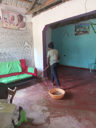 An interior scene within a residence in Manda-Shivanga ward, Kenya, depicts an individual actively cleaning a dirt and water-covered floor. A dark-skinned person, wearing a dark beanie, light olive-green t-shirt, and dark trousers, stands barefoot, facing away from the camera, and uses a stick or broom to push murky, brown water and dirt across the reddish-brown floor. An orange plastic basin containing dirty water is positioned on the floor in front of this individual. In the immediate left foreground, the back of another dark-skinned person's head and shoulder are partially visible; this individual is seated on a wooden chair, wearing a striped shirt, with a green fabric on their lap. The floor throughout the visible area is extensively covered with wet dirt, mud, and standing water. To the left, a sofa with a red base and green fabric covers is situated against a textured white wall adorned with a large framed picture depicting a praying family. The ceiling consists of exposed wooden beams supporting a corrugated metal roof. An archway with dark purple trim separates the foreground from a back room with light blue/teal walls, which displays a framed image of a green-roofed building and a green garland.