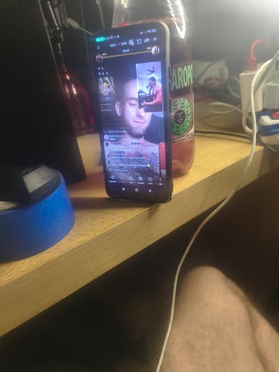 A close-up, high-angle shot captures a smartphone displaying a live video stream, placed on a wooden desk. The phone, oriented vertically, shows a split-screen view: the left side displays a smaller profile of a person with a tattoo on their arm, while the right side features a larger view of a shirtless man with a goatee and a headband. Chat messages are visible below the video, including one that reads "Obsidian" and "Battle Results: Draw." To the left of the phone, a blue and black object, possibly a mouse or a device accessory, is taped with blue tape. In the background, a partially visible bottle with a brown liquid and a label containing nutritional information stands to the right of the phone. A white cable snakes across the desk and leads to a white power strip with multiple plugs. The overall environment suggests an indoor setting, likely a workspace or personal area. The image does not provide any information that would allow for the determination of the location in the city of Grójec, Poland.