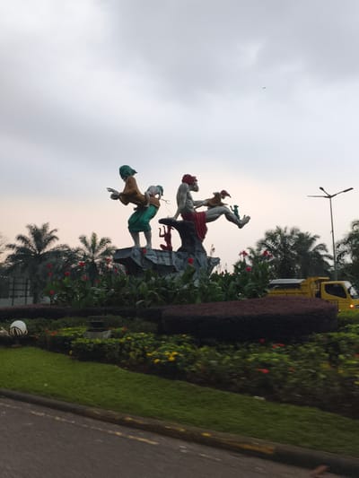 The image displays a vibrant, multi-figure statue situated within a well-maintained roundabout or landscaped median. The central artwork features two primary figures perched on a grey rock formation. On the left, a woman with green hair and draped in green and ochre clothing carries a small child on her back. To her right, a man with reddish hair and a red lower garment sits, engaging with a smaller, brown animal-like creature held on his lap, while a green animal-like figure rests near his feet.
The statue is surrounded by lush landscaping, including neatly trimmed green grass, dark green and deep red hedges, and clusters of bright red flowers, possibly Canna lilies. Numerous palm trees are visible in the background, hinting at a tropical setting. On the far right, a yellow dump truck and a tall street light pole are partially obscured by the foliage. The foreground shows a section of a road with a visible curb and faint yellow road markings.
The sky is overcast and cloudy, but a soft, warm glow near the horizon suggests either early morning or late afternoon light. A tiny bird can be seen flying in the upper sky. The scene is quiet and orderly, showcasing public art in an outdoor environment, consistent with a location like Babakan Madang, Indonesia. No discernible text is visible in the image.