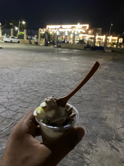 A hand holds a disposable plastic cup filled with ice cream and a wooden spoon inserted into it, on a cobblestone pavement at night. In the background, a white car is parked near a building with bright lights and a fence. Several people are visible walking and standing near the illuminated building, which appears to be a restaurant or marketplace. The scene is likely located in Tipaza, Algeria.