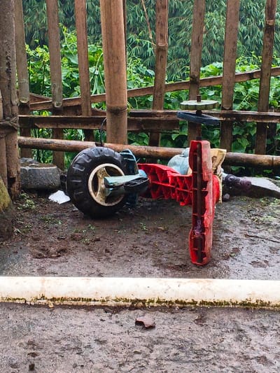 A red and black toy tricycle sits in muddy ground. Its black tire and worn red plastic parts are visible. Behind it, a crude bamboo fence provides a backdrop. A white pipe lies on the lower portion of the frame. The ground is wet and textured. The background greenery is dense and blurred.