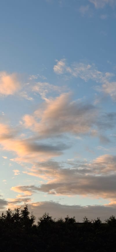 The image depicts a twilight sky over a silhouette of trees, likely taken in Orimattila, Finland. The sky is a soft blue, transitioning to hues of pink and orange where the clouds are catching the last rays of sunlight. The clouds are scattered, some appearing wispy and high, while others are denser and lower, casting a slight greyish tone in places. The trees at the bottom of the image form a dark, irregular line against the brightly lit sky, their shapes indistinct due to the low light. There are no people or specific man-made structures clearly visible, suggesting a natural landscape scene.
