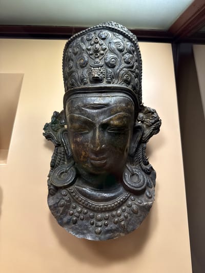 The image shows a bronze sculpture of a Buddha head. The sculpture is detailed, with intricate designs on the headdress and around the face. It is mounted on a light beige-colored wall. The sculpture appears to be indoors, possibly in a museum or a display case, evident from the window frame visible on the left side of the image. It is speculated that the sculpture may be located in the city of Lalitpur, Nepal.