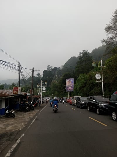 The image shows a street in the Mae Chan District, Thailand. The sky is overcast. The road is congested with a line of cars and motorcycles moving slowly. Some motorcycles are parked on the side of the road, near a small shop with signage in an Asian language. Power lines are visible above the shop. The road is surrounded by lush green vegetation and trees on the right, sloping uphill. Signposts and advertising billboards are interspersed along the roadside.