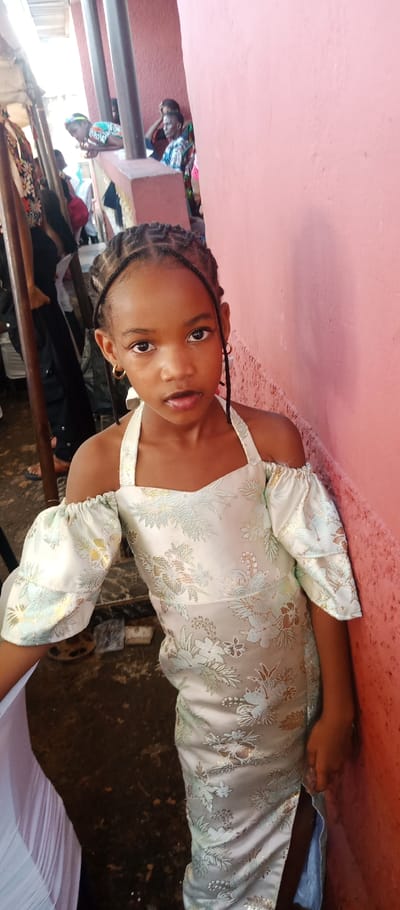 A young girl, approximately 6-10 years old, stands in the city of Udi, Nigeria, positioned slightly right of center, gazing directly forward. She has dark skin and her hair is styled in dark cornrow braids. Her lips are slightly parted, and she wears small gold-colored hoop earrings. She is dressed in a light-colored, possibly pale mint green or off-white, floor-length gown featuring a floral pattern in light blue/green and metallic gold thread. The dress has a halter neckline, off-the-shoulder puffed sleeves with elasticized cuffs, and a visible thigh-high slit on the lower right side. Her right arm is pressed against a textured, plain pink wall that occupies the right half of the image.
To the left of the girl, the environment is open and occupied by several individuals and structural elements. A person in dark attire (black top and pants) is partially visible in the mid-ground left. Further back, two dark brown vertical posts support an overhead structure, likely a light-colored corrugated roof or ceiling. Behind these posts, multiple individuals are discernible, some standing and others leaning on a low brown concrete railing. One woman wears a colorful patterned shirt while leaning on this railing. The ground in the foreground and mid-ground is dark, uneven, and appears to be earth or concrete, with some scattered debris. White pleated fabric is visible on the far left, adjacent to the girl's left side. The scene is illuminated by bright, diffused light, indicating a daytime, semi-outdoor setting, possibly during an event.