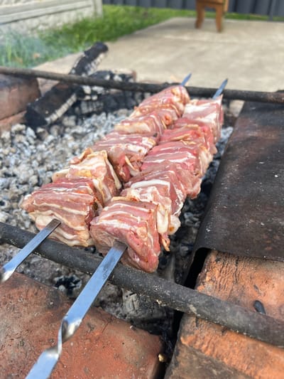 Two skewers of raw, cubed meat, likely pork or lamb, are positioned over a bed of smoldering charcoal within a makeshift brick grill. The meat appears to be seasoned with visible specks of spices. The grill itself is constructed from rough, reddish-brown bricks. In the background, a concrete patio surface is visible, with a wooden structure and some green vegetation beyond. The scene is outdoors, suggesting a barbecue or outdoor cooking event in the village of Buchatsin, in the Buchatsin rural council of Belarus. There are no people visible in the image.