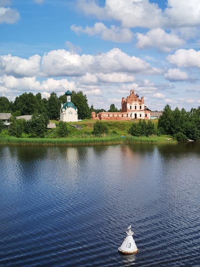 This is a medium-shot, eye-level, vertical, outdoor, daytime, landscape photograph taken from a body of water looking towards the shore in Anchimovo, Russia. The image features a calm, dark blue river in the foreground, with ripples reflecting the sky. In the mid-ground, the shoreline is lined with tall green reeds and grass. Behind the reeds, there are trees and two buildings. On the left, a white chapel with a green dome stands prominently. To its right and slightly further back, a ruined brick building, likely an old church, is visible. The background consists of lush green trees and a bright blue sky filled with fluffy white clouds. A white buoy with the number "19" is floating in the river in the lower right quadrant of the image.