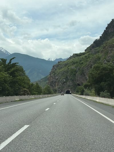 An outdoor, eye-level, medium shot shows a car driving down a highway towards a mountain tunnel. The highway is asphalt, with white painted lines dividing the lanes. The highway is flanked by concrete barriers, beyond which is green grass and trees. The mountains are covered in greenery, with a few snow-capped peaks visible in the distance. The sky is overcast with white clouds. The car is a light-colored SUV, and it is the only vehicle visible on the highway. The tunnel is dark and appears to be a large, man-made structure. The scene is peaceful and scenic. The image does not show any people.