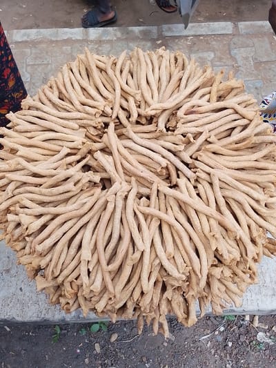 A pile of fried snacks, likely of the "kuli kuli" variety, is displayed on a concrete surface. The snacks are long, thin, and twisted, with a golden-brown, slightly irregular texture. They are arranged in a circular mound, with many individual pieces overlapping and creating a dense mass. The lighting suggests it is daytime. In the background, the feet of at least one person are visible, wearing dark sandals. A portion of patterned fabric, possibly clothing or a textile being sold, is also visible on the left side of the image. The ground beneath the concrete is a mixture of dirt and small stones. The scene appears to be an outdoor market setting, possibly in Oja Oba, Oko Erin, Nigeria. There is no visible text in the image.