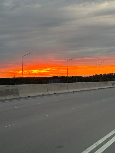 The image shows a highway adjacent to a treeline with a vivid sunset sky. The highway is gray asphalt with painted white lane markings. A concrete barrier separates the highway from the trees. Streetlights are evenly spaced along the treeline. The sunset is primarily orange and yellow. The sky on the opposite side of the sunset is a cloudy gray. The location is most likely in the city of Avdon, Russia, based on the prompt.