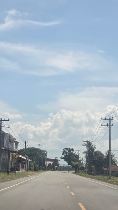 The image shows a road in Jurong Pandee, Indonesia. The road is paved and has yellow dashed lines. Utility poles with wires run along the side of the road, near some trees and houses. The sky is visible with white clouds.