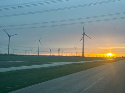 The image shows a landscape with several wind turbines. The sun is setting in the background, creating a bright orange glow on the horizon. In the foreground, there is a road and green fields. High-voltage power lines are visible above the landscape. This image captures a scene in Lelystad, Netherlands.
