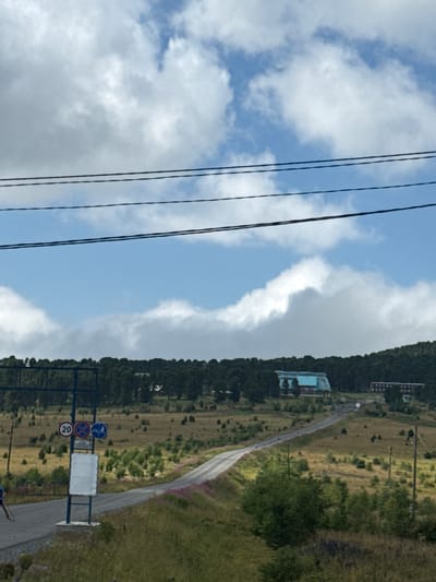 The image depicts a rural landscape under a cloudy sky. In the foreground, a road winds through rolling hills covered in dry grass and scattered bushes. A figure on skis is visible on the left side of the road, near a blue metal signpost with traffic signs and a blank white panel. The road leads towards a distant forested hillside. On the hillside, two buildings are visible: a larger, light-blue structure with a distinctive curved roof, and a smaller, darker rectangular building. A series of power lines stretches across the upper portion of the image. The overall impression is one of a remote, possibly mountainous, area.