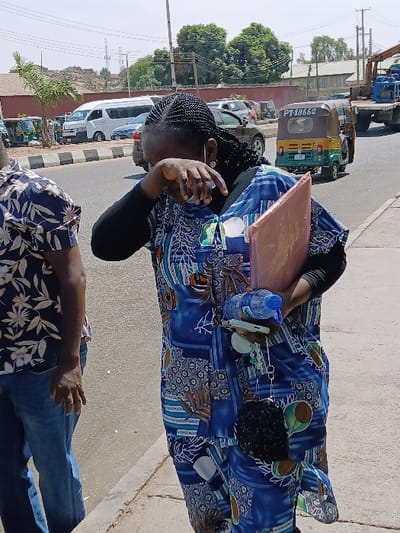 The scene depicts an outdoor street in Communauté rurale de Payar, Senegal, under clear daylight. A woman, appearing to be of African descent, occupies the right foreground. She wears a blue and multi-patterned kaftan over a black long-sleeved garment, with her dark braided hair visible. Her right hand is raised to her face, partially obscuring her eyes. Her left hand carries a clear plastic water bottle with a blue cap, a light-colored smartphone, a key chain with a black fuzzy ornament, and a reddish-brown folder. A person, partially visible on the left, wears a blue floral-patterned shirt and blue jeans. The background features an asphalt road bordered by a concrete curb. Vehicles on the road include a white passenger van, other cars, a yellow and green auto-rickshaw displaying registration 'PT-18662', and a flatbed truck with blue machinery. A small palm tree and green deciduous trees are present, along with overhead power lines and a distant red-brown wall. Rocky outcrops are visible in the far left background.