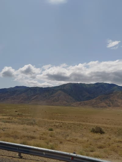 The image captures a vast, arid landscape with a mountain range in the background. The sky is partially cloudy with patches of blue. The foreground features a metal guardrail and a dry, grassy plain dotted with sparse vegetation. The mountains themselves are rugged and appear to be covered in a mix of brown and green, suggesting dry brush and some hardy vegetation. The overall impression is one of a remote, undeveloped area. The scene is consistent with the geography found in and around Ынталы, Kazakhstan, a region characterized by its steppe and mountainous terrain. There are no people or specific structures visible in the image.