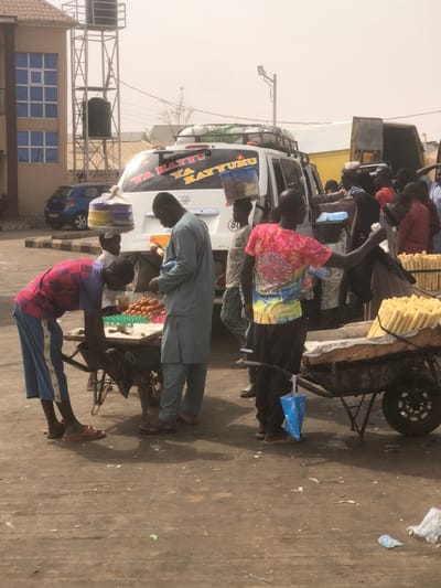 The scene depicts a bustling street market in Unguwar Fatika, Nigeria, under a hazy sky. In the foreground, two mobile vendor carts are present on a dirt and concrete surface. On the left, a male wearing a red and blue plaid shirt and blue shorts is bent over a wooden cart displaying rows of brown eggs in green and white plastic cartons. Adjacent to him, a male in a light blue traditional tunic and trousers stands upright, looking down at the cart's contents. Behind them, a white passenger van, heavily loaded with luggage on its roof rack, features "YA HAYYU YA KAYYUMU" in red script on its rear window. A young male balances a large, multi-colored, wrapped item on his head while standing near the van's rear. Further right, another male in a tie-dye shirt (red, yellow, blue) stands next to a second wooden cart filled with numerous peeled sugarcane sticks. He holds a clear plastic bottle in his left hand, with a blue plastic bag hanging from his right wrist. A cluster of individuals, primarily male, are gathered further right in the mid-ground, near a partially visible yellow truck. In the background left, a multi-story building with blue-tinted windows is visible, along with a metal structure supporting a dark water tank. A blue car is parked behind the building. The ground shows scattered debris, including plastic bags.