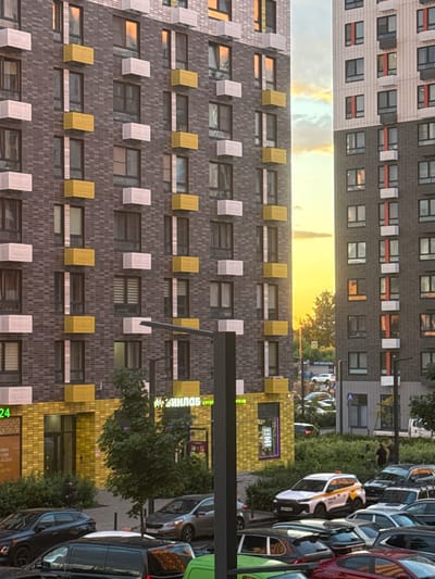 The media file displays a street scene in Drozhzhino, Russia, characterized by modern apartment buildings and a variety of parked cars. The perspective is somewhat distorted, giving the impression of a tall, multi-story structure filling the frame. Several cars are visible, including a distinctive white and yellow car that appears to be a taxi, with visible text on its side. Other vehicles include a red car, a green car, and several darker colored cars, some of which are partially obscured.
The apartment buildings feature a patterned brick facade with contrasting architectural elements such as white and yellow rectangular protrusions above some windows. The windows themselves are dark, reflecting the surroundings. A bright, sunlit sky with scattered clouds suggests it is daytime, likely late afternoon or early evening given the warm hues of the sunlight. There are also areas of dense green foliage, indicating the presence of trees and bushes, possibly in a park or courtyard area adjacent to the buildings and parking lot.
On one of the buildings, a yellow storefront with a glass entrance is visible. Above the entrance, illuminated green text in Cyrillic characters can be seen. Some of this text appears to read "ЦИФРЛАВ СУПЕР". Another sign with green text is partially visible near the bottom of the frame, with the number "24" prominent. The overall scene depicts an urban residential environment with elements of everyday life, including transportation and commercial activity.
