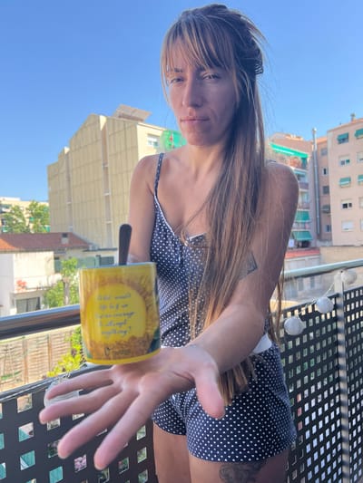 A woman with long, light brown hair stands on a balcony, holding a yellow mug with a sun design and text. The mug has a black spoon inside. She wears a dark blue pajama set with white polka dots and has a tattoo on her left arm. The background shows buildings and a clear blue sky, suggesting an urban setting. The balcony has a black metal railing with decorative white string lights. The image appears to have been taken in Mollet Del Vallès, Spain, given the architectural style of the buildings.