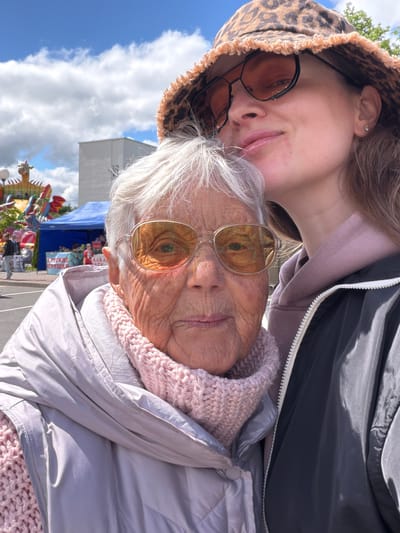 Two people pose for a selfie. An older woman with gray hair, wearing glasses and a pink scarf under a light gray jacket, looks directly at the camera. A younger person, wearing a leopard print hat and dark-rimmed glasses, presses their face close to the older woman's. The background shows a partly cloudy sky, a white building, and colorful fairground attractions, including tents and figures. The location is likely a public space in Valozhyn, Belarus, given the Cyrillic lettering visible on the tents.