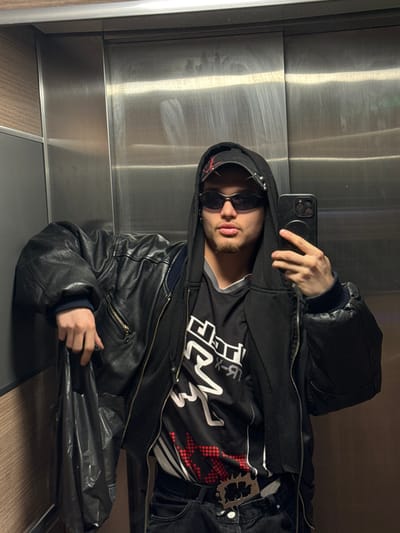 The image captures a single male individual taking a selfie inside an elevator in Asnières-sur-Seine, France. The subject, appearing to be in his late teens or early twenties, has short facial hair on his chin and wears dark, oval-shaped sunglasses. He is dressed in a black backward baseball cap with red detailing and metal studs, a black hooded sweatshirt with the hood up, a black leather bomber jacket, and a black t-shirt with a white and red graphic design. He also wears dark trousers with a prominent metallic belt buckle featuring a jagged outline and intricate design. In his right hand, he holds a black smartphone, positioned to capture the image. His left hand holds a dark, crumpled plastic bag, resting against his left side. The elevator interior consists of brushed metallic panels, exhibiting vertical streaks and reflections, with a narrow vertical panel of light wood-grain material visible on the left.