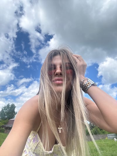 The image is a selfie taken outdoors, likely in a rural area, with a woman in the foreground. She has long blonde hair and is wearing pink-tinted sunglasses and a light-colored dress with a floral pattern. Her left hand is raised and touching her head. She is wearing a watch on her left wrist. In the background, there is a cloudy blue sky, green grass, trees, and a wooden structure. The location is specified as Пастухово, Russia.