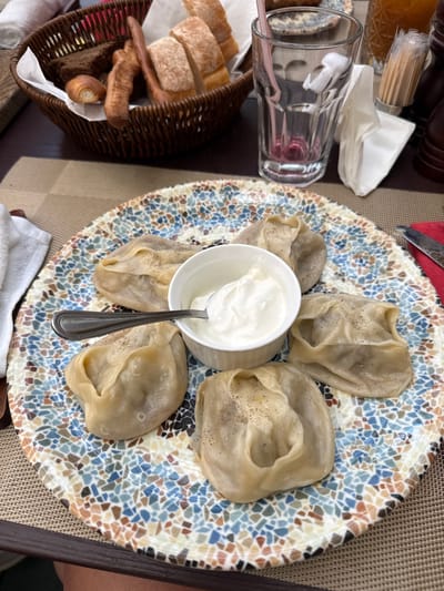 The image shows a close-up, overhead view of a plate of manti, a type of dumpling, served with a dollop of sour cream in a small white bowl. The manti are arranged around the bowl on a decorative plate with a mosaic-like pattern of blue, brown, and white tiles. A spoon rests in the sour cream. To the left of the plate, there is a woven basket filled with bread, including sliced baguettes and smaller rolls. A clear glass with a straw and some pink liquid is also visible, along with a packet of toothpicks and a white napkin. The setting appears to be a restaurant or dining area, with a textured placemat on a table. The lighting suggests it might be daytime. The focus is on the food, with no people fully visible, though a hint of a person's leg is visible on the right side of the frame. The scene is likely in Zhukovka, Russia, as indicated by the location context. There is no visible text in the image.