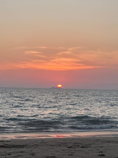 The image depicts a sunset over a body of water at Phra Ae beach in Phra Ae, Thailand. The sky exhibits a gradient of colors, transitioning from light peach and pale orange at the top to a vibrant orange and red hue near the horizon. Elongated, wispy clouds are present, primarily in the middle section of the sky.
The sun, a distinct orange-red circular orb, is partially visible and appears to be setting behind a low, dark landmass on the distant horizon. This landmass forms a silhouette against the bright sun.
Below the horizon, the water's surface is a dark blue-grey, characterized by ripples and small breaking waves. A faint, reddish reflection of the sun is visible on the water closer to the foreground. A small, dark, circular object, possibly a buoy, is discernible floating on the water in the mid-ground to the left of the frame.
The foreground consists of a sandy beach, appearing dark brown-grey. The sand shows variations in texture and color, indicating both dry and wet areas, with the wet sand closer to the water's edge. Minor indentations or marks are visible on the sand. No people, major objects, or significant actions are depicted in the scene.