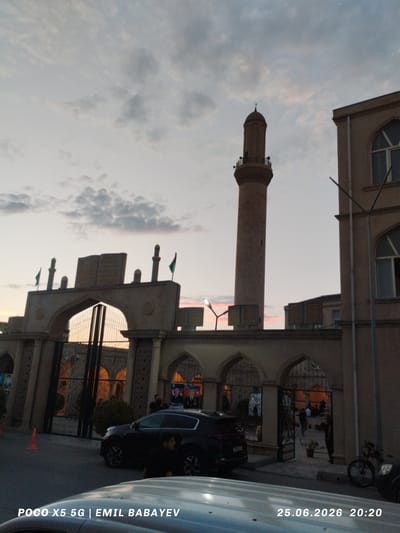 A photograph captured at dusk shows the exterior of a mosque complex in Sabunchu Raion, Azerbaijan. The sky is filled with soft clouds, tinged with the colors of sunset, suggesting it is early evening. In the foreground, the tops of two vehicles are visible.

The main focus of the image is a large, ornate entrance gate with arched openings. Beyond the gate, an open courtyard is visible, with several people walking and standing. A tall, slender minaret rises prominently from behind the mosque buildings, its silhouette sharp against the fading light. The architecture of the complex features Islamic design elements, including intricate carvings and decorative motifs. Flags can be seen atop the entrance gate.

On the right side of the image, part of a building with arched windows is visible. A street lamp with a single light shines. A bicycle is parked near the right edge of the frame.

The overall mood is peaceful and serene, typical of a quiet evening in a religious complex. The lighting suggests the transition from day to night.

Visible text in the image includes "POCO X5 5G | EMIL BABAYEV" in the lower left corner and "25.06.2026 20:20" in the lower right corner.