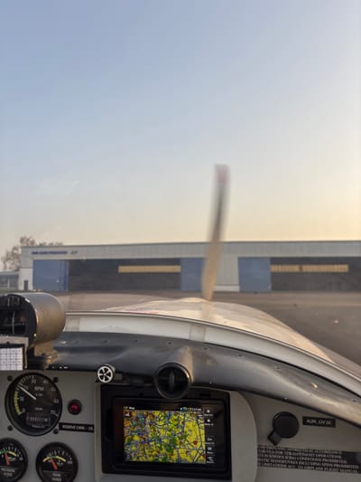 The image presents a first-person view from the cockpit of a light aircraft on the ground at Toussus-le-Noble, France. In the immediate foreground, a portion of the aircraft's instrument panel is visible. Key instruments include an RPM gauge indicating approximately 1000 RPM, an hours counter, and a functional GPS navigation screen displaying a detailed geographical map with roads and land features. Labels for "RESERVE CARB. - 10L," "PRESSION HUILE" (Oil Pressure), "TEMP HUILE" (Oil Temperature), and "ALIM. 12V DC" are partially discernible. A small white knob and an air vent are also present on the panel.

Through the aircraft's windscreen, the propeller is visible in the midground, significantly blurred, indicating active rotation. The windscreen shows minor surface imperfections. Beyond the aircraft, an asphalt or paved airfield surface extends to two large hangars in the background. These hangars feature grey facades with prominent blue sliding doors. The hangar on the left displays the text "AIR HOP FORMATION" and a partial identifier, possibly "F7." A sparse line of trees is visible behind this hangar. The sky above is clear, exhibiting a gradient from light blue at the zenith to a pale yellowish-orange hue near the horizon, consistent with low sun angles during sunrise or sunset. No individuals are depicted within the frame.