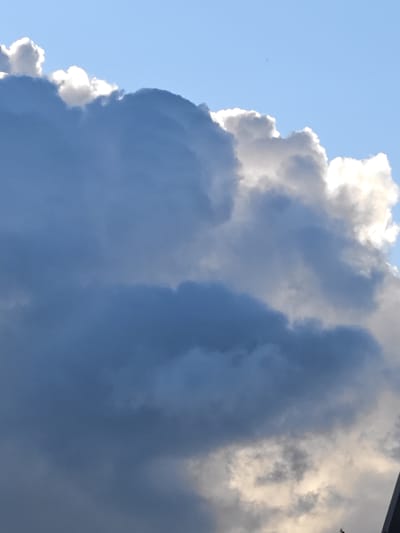 A low-angle, medium shot captures a dynamic sky with dramatic clouds and a clear blue sky. The clouds are a mix of dark grays and bright whites, with the sunlight dramatically backlighting some of the cloud formations, creating a luminous effect. The clouds appear to be large and dense, suggesting potential for rain or stormy weather. The lighting indicates it is daytime, likely late afternoon or early evening given the quality of light. No people, animals, or man-made structures are clearly visible in the frame, except for a sliver of a dark, possibly building structure, in the lower right corner. The location is identified as Rodniki, Russia.
