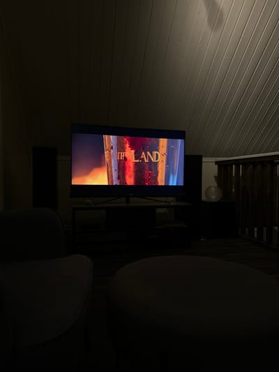 A dimly lit room showcases a television screen displaying the words "The Land," oriented sideways. To the right, a chair's silhouette dominates the scene, its design barely discernible in the low light. Above the television, a dark structure with slats is visible, possibly a deck or staircase. A small, spherical object hangs nearby. The left side of the image features light-colored siding, adding contrast to the overall darkness of the setting.
