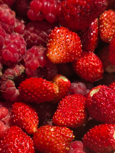 A close-up, overhead shot displays a vibrant pile of red raspberries and small wild strawberries. The fruits are closely packed together, with the textures of the raspberries' drupelets and the strawberries' seeds clearly visible. The lighting emphasizes the glossy surfaces of the berries, creating highlights and shadows. The scene is static, with no discernible actions or interactions. The location is identified as Восход Снт (фролово), Russia, implying an outdoor setting, likely a garden or market stall, given the subject matter. The image is focused on the natural beauty and texture of the fresh produce.