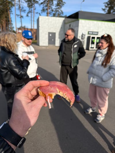 The image appears to be taken from a first-person perspective, focusing on a partially eaten sandwich held in a hand wearing a black watch. The sandwich consists of bread topped with a slice of meat, possibly ham or prosciutto, and a small amount of white spread, potentially mayonnaise or cream cheese. In the blurred background are three individuals. One is wearing a white puffer jacket and light pink pants, another wears a dark jacket with a green vest and black pants, and the third wears a white jacket, a black leather jacket, and a blue cap. The group seems to be standing near a building, possibly a shop or a station. Given the description in the prompt, the location is presumed to be in Luga, Russia. The time of day appears to be daytime. There is no clear indication of any particular actions, interactions, or specific environmental details, other than the building and a few trees visible in the upper portion of the image.