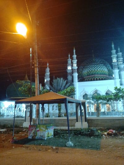 The image depicts a night scene in Kotapantonlabu, Indonesia, featuring a mosque with multiple minarets and a central dome illuminated by artificial lighting. In the foreground stands a simple structure, likely a vendor's stall, with advertisements visible. The environment includes a roadside area with visible ground clutter. A streetlamp illuminates the scene, casting a warm glow. No people are visible in the image.