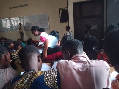 A classroom scene shows many students sitting at desks, facing forward. A female student with a white and pink shirt is seen near the front, leaning over papers. A whiteboard with writing on it is visible in the background, along with a man standing near it. More individuals stand or walk near a doorway at the back of the room. The overall atmosphere appears to be academic.