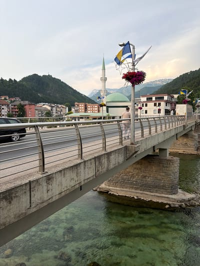 The image depicts the Old Bridge in Konjic, Bosnia and Herzegovina, spanning across the turquoise Neretva River. The bridge, constructed from stone and concrete, has a modern railing system. On the bridge, a person dressed in light-colored clothing is walking away from the camera. A black car is visible in the left lane of the bridge, and a truck is parked in the right lane.
In the background, a minaret and a green dome of a mosque are prominent architectural features. The city's buildings, nestled against lush green mountains, form a picturesque urban landscape. The sky is overcast, with hints of sunlight breaking through the clouds. The clear, shallow water of the Neretva River reveals rocks and pebbles below the surface. Flags are visible on poles along the bridge, suggesting a celebratory or national occasion.