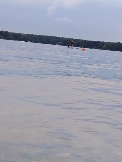 The image depicts a wide shot of a body of water, likely a reservoir, with a treeline in the background and a cloudy sky above. The water's surface is rippled, creating patterns of light and shadow. In the middle ground, two individuals are visible on stand-up paddleboards. One person, wearing an orange life vest, is standing and appears to be paddling. Another person in a similar vest is also on a paddleboard, further in the distance. An orange buoy is floating in the water between the paddleboarders. The overall scene suggests a recreational activity taking place on a lake or reservoir, possibly during the daytime given the visible light. The overcast sky indicates cloudy weather. The treeline suggests a natural, possibly forested, environment surrounding the water. There is no visible text in the image.