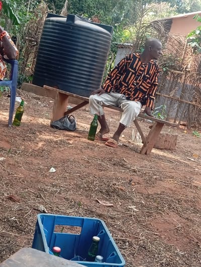 The image depicts an outdoor scene in Egede, Nigeria, characterized by reddish-brown, dry earth with scattered organic debris. A bald, dark-skinned male subject, wearing a black and orange patterned long-sleeved shirt and light-colored ripped trousers, is seated on a simple wooden bench. He wears one brown sandal on his right foot and is looking towards the right side of the frame. To his left, a partial view of another individual shows an arm with a red beaded bracelet, seated on a blue plastic chair.

Behind the seated man, a large, black, ribbed plastic water tank is elevated on a wooden support structure. On the ground near the man's feet, a green glass bottle without a cap is visible, and another similar bottle is positioned near the blue chair on the far left. A black plastic bag rests on the ground under the wooden bench. In the immediate foreground, a blue plastic crate contains at least three capped bottles: one green glass bottle with a yellow cap, another with a red cap, and a third partially visible.

The background features sparse vegetation, including dry leaves and taller plants, with a light-colored building or structure visible on the upper right side. The scene is illuminated by natural daylight.