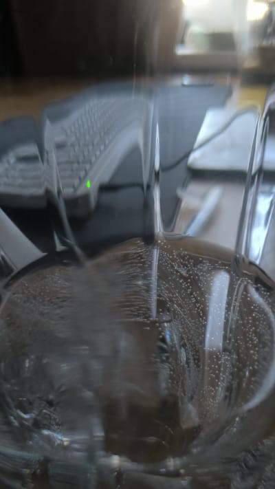 A close-up, out-of-focus shot captures a glass with a carbonated beverage, likely sparkling water or soda, as indicated by the numerous small bubbles rising to the surface. The drink appears to be clear and is contained within a transparent glass. In the background, a keyboard is visible, suggesting the setting is likely a desk or workspace. A faint green light on the keyboard indicates a device is active. The lighting is diffuse, and the overall image has a shallow depth of field, with the foreground bubbles and the glass being the primary subjects. The scene does not provide any specific geographical or contextual information that would allow for an identification of the city of Kovrov, Russia. There are no people or discernible interactions present in the image.