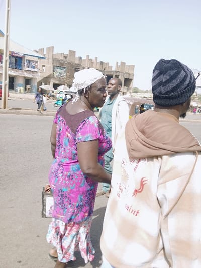 The image depicts a street scene in Mumbwa, Zambia, under clear daylight conditions.
In the immediate foreground, to the right, a person with dark skin is seen from the back and right side. This individual wears a black and gray striped knitted beanie and a light brown hooded sweatshirt with a white graphic on the back, featuring a stylized animal figure with red accents and the word "saint" in white. A dark beard is visible.
To the left of the foreground, an adult woman with dark skin is shown in profile, facing left. She wears a white, possibly crocheted or beaded, head covering. Her attire consists of a short-sleeved top with a dark purple upper section and a vibrant pink, blue, and black floral or abstract pattern, paired with a light-colored, possibly white and pink, floral patterned skirt with a ruffled hem. She holds a dark object in her left hand.
Further back, between the two foreground subjects, a man with dark skin is partially visible, oriented towards the right. He wears a light-colored long-sleeved shirt over a green or teal undershirt.
The subjects are standing on a paved surface.
In the background, on the left, a two-story building with a light-colored facade and blue window frames is visible, appearing to house shops on the ground floor. Across the street, a large, multi-story concrete structure, possibly unfinished or in disrepair, dominates the view, characterized by angular forms and exposed construction elements. A large poster or mural is visible on one section of this structure. The street also features several pedestrians and multiple three-wheeled vehicles, resembling tuk-tuks. The sky is clear, indicating bright weather. No explicit interaction is observed between the foreground individuals; they appear to be present in a public thoroughfare.