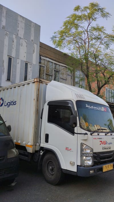A white Isuzu ELF logistics truck is parked on an asphalt road in front of a multi-story building with a distressed facade. The building features vertical grey and white stripes on the left side, and boarded-up windows on the upper floors to the right. A large tree with green foliage grows to the right of the building, partially obscuring the sky. The truck has "Logistics" written in blue on its container and "ISUZU ELF" on its fender. Arabic script is visible on the windshield, along with the license plate "B 9496 TXV". Another vehicle, partially visible and black, is parked to the left of the truck. The lighting suggests it is daytime, likely late afternoon given the long shadows. The scene appears to be located in the city of Trosobo, Indonesia, as indicated by the license plate format.