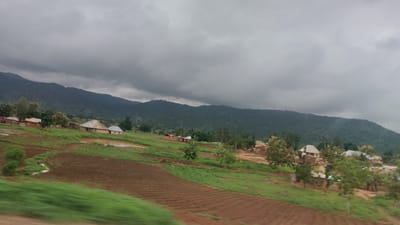 The image depicts a rural landscape in Gidan Waya, Nigeria, under a heavy, overcast sky. Rolling hills covered in dense, green vegetation dominate the background, with a range of mountains stretching across the horizon. In the mid-ground, a small cluster of traditional houses with corrugated metal roofs are nestled on a hillside. Some of these houses appear to be made of mud or clay.

In the foreground, cultivated fields with rich brown soil are visible, some showing clear rows of tilled land, suggesting agricultural activity. Patches of vibrant green grass and smaller trees are interspersed throughout the scene. A small, calm body of water, possibly a pond or a flooded area, is situated near the houses. The overall impression is one of a serene, rural community situated amidst a lush, natural environment. There are no people or animals visible in the image.