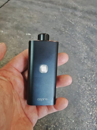A hand holds a dark gray electronic cigarette device, likely made of metal, against a rough, textured concrete surface. The device has a small, square, illuminated button in the center and the word "aspire" is visible in white lettering on the lower portion of the device. The concrete appears to be an outdoor surface, possibly a sidewalk or patio, with visible aggregate. The image's lighting suggests it was taken during the daytime, possibly outdoors. The exact location in Hod HaSharon, Israel, is not discernible from the visual information provided in the image.